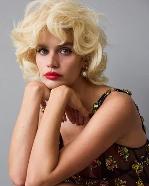 Model Sara Sampaio with blonde hair and red lipstick wearing a dark embellished dress against a plain grey background