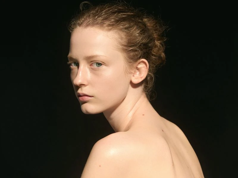 Portrait of a fair-skinned young woman with green eyes and curly auburn hair pulled back, set against a dark background in a studio.