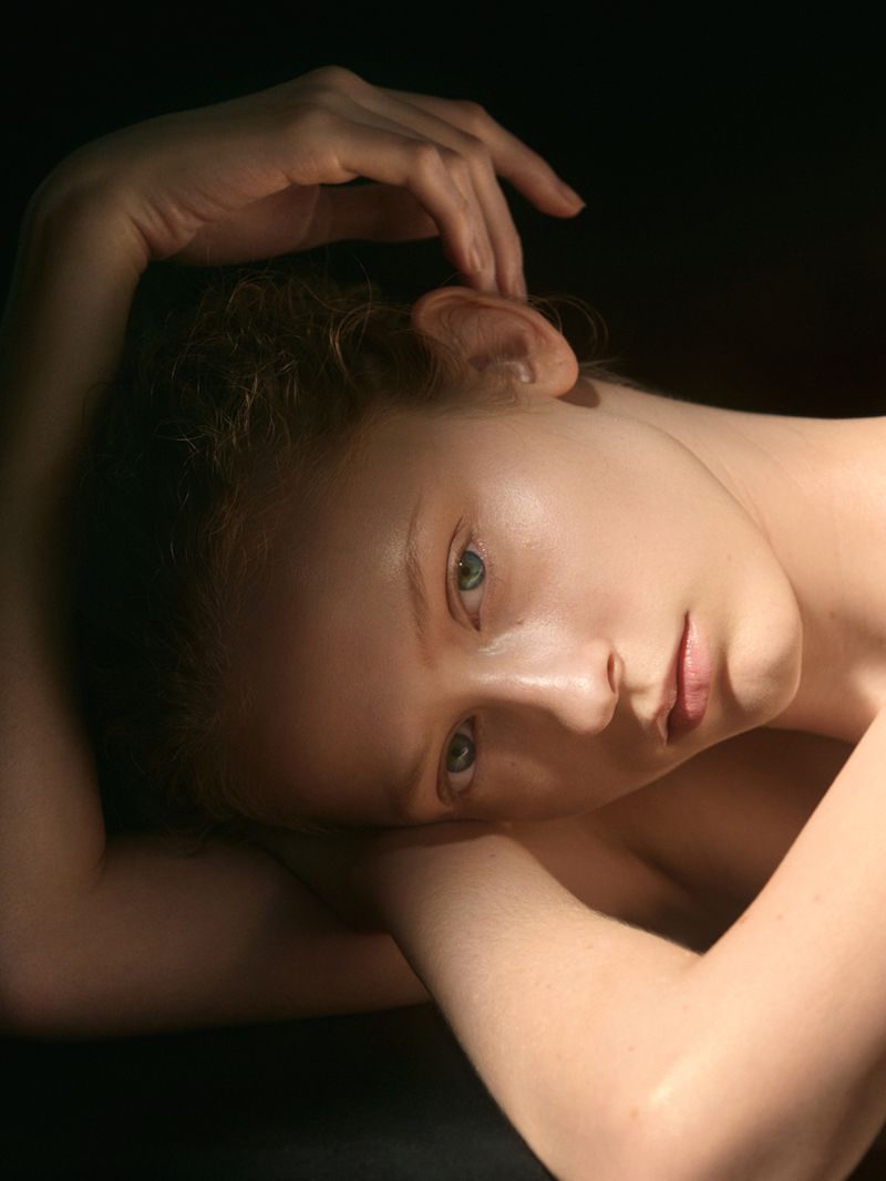 Close-up portrait of a young woman with fair skin and light hair, lying down with her arms above her head, against a dark background.