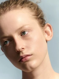 Close-up beauty portrait of a female model with natural skin and tied-back hair against a solid light blue background.