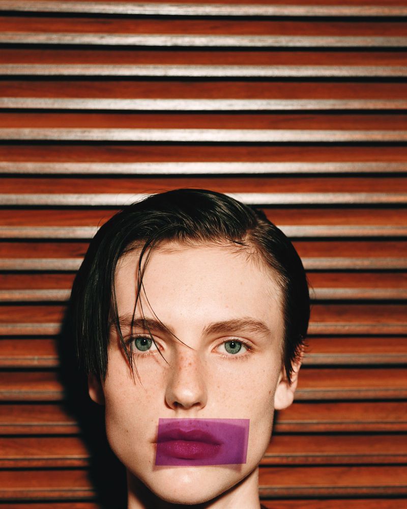 Close-up portrait of a person with blue eyes and dark hair, featuring a purple rectangle covering their lips, against a wood-slat background.