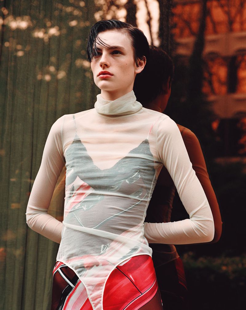Fashionable woman poses in sheer top and patterned bottoms with soft window light and blurry background reflection, eye-level shot.