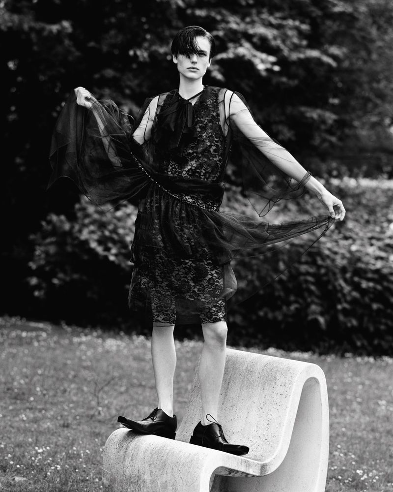 A person standing on a bench outdoors wearing a black lace dress, posing in a black and white fashion photograph with trees in the background.