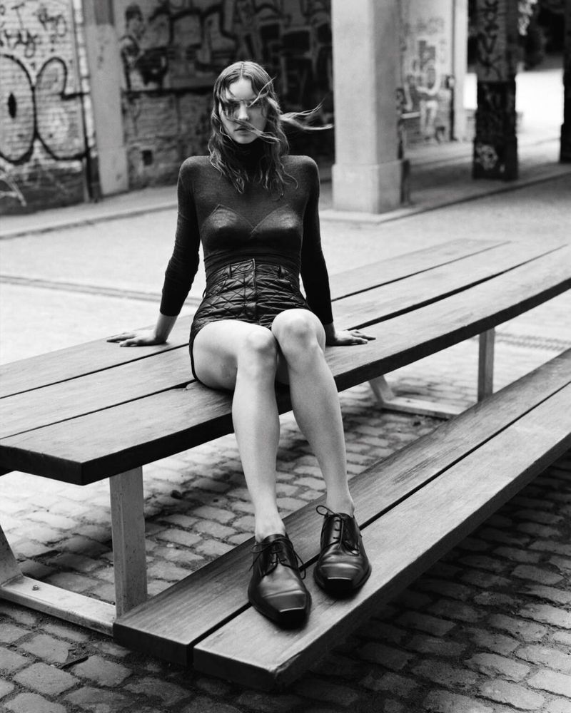 Monochrome portrait of a woman in black top and shorts sitting on a wooden bench, wearing black shoes, against a graffiti-covered wall.