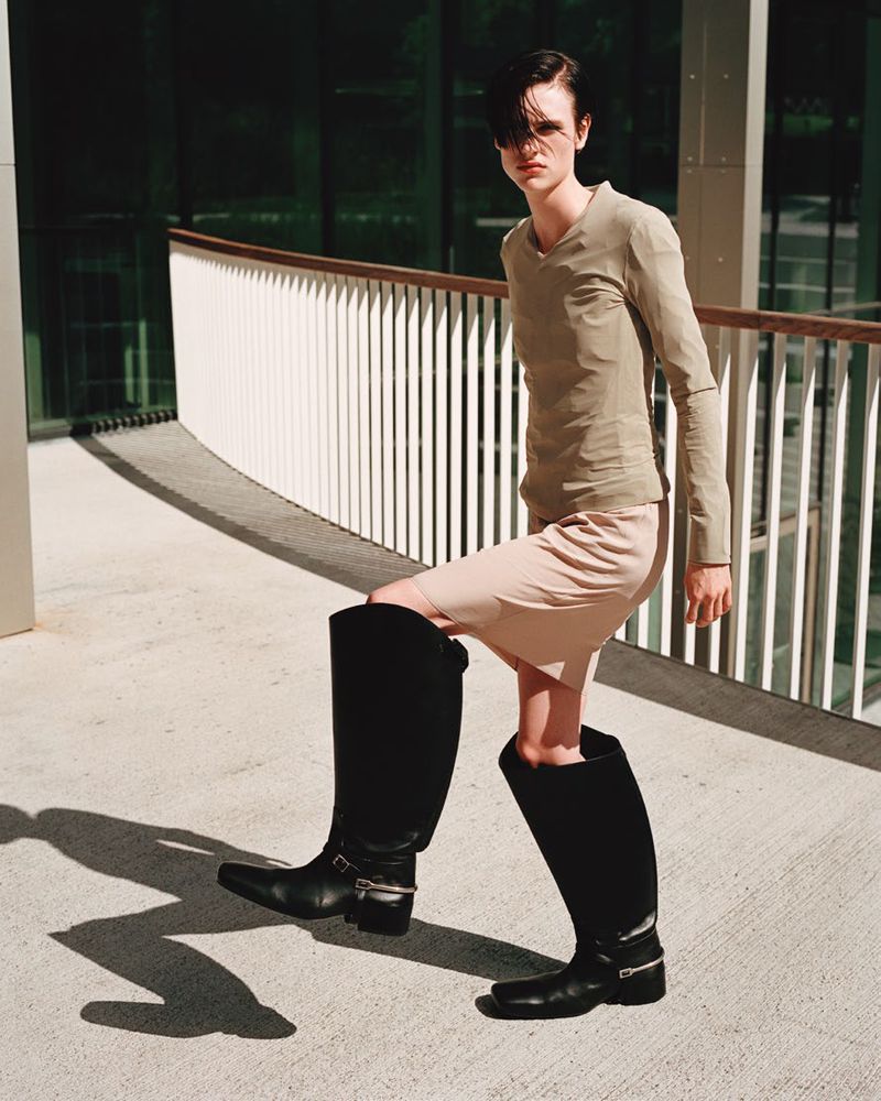 A slender adult stands outdoors wearing black leather riding boots, a khaki-colored shirt, and a light pink skirt with a neutral facial expression.
