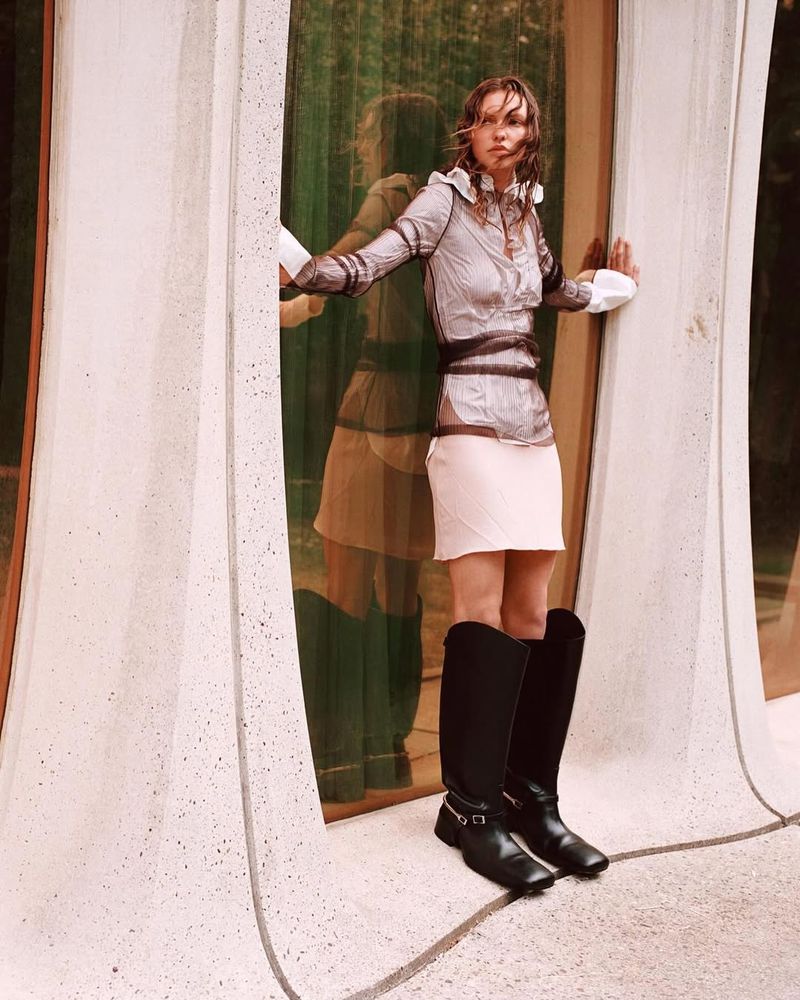 A young woman poses in a translucent top, mini skirt, and tall boots, standing in a modern doorway with reflected light. Fashion shot.