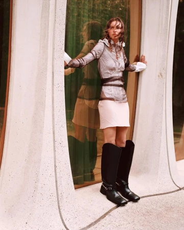 A young woman poses in a translucent top, mini skirt, and tall boots, standing in a modern doorway with reflected light. Fashion shot.