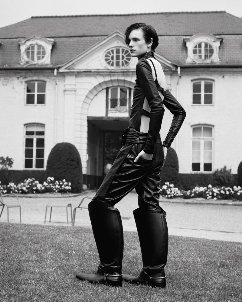 Monochrome fashion shot of a slender adult in dark leather overalls and tall boots posing outdoors with a large house in the background.