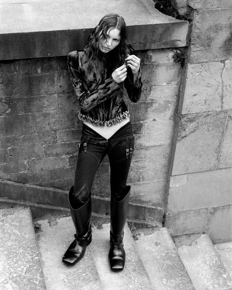 A stylish model poses on stone stairs in a black velvet jacket, dark jeans, and high black boots in a black-and-white fashion shot.