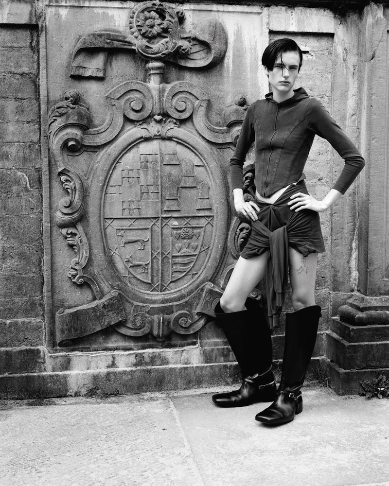 Monochrome outdoor fashion shot of a young adult woman in modern gothic fashion with knee-high boots, posing near a stone wall.