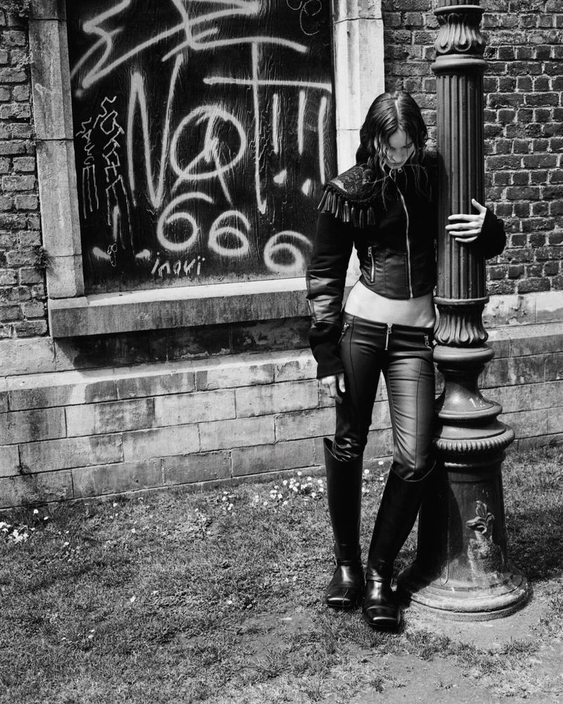 A woman with long hair leans against a pillar in an urban environment. She is wearing a jacket, jeans and boots. There is graffiti in the background.
