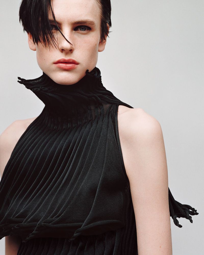 Fashion portrait of an androgynous model in a black, high-necked, pleated dress against a plain background in a studio setting.