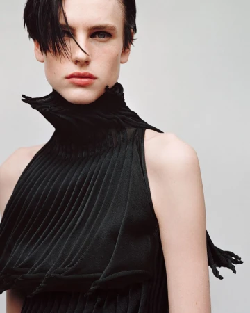 Fashion portrait of an androgynous model in a black, high-necked, pleated dress against a plain background in a studio setting.