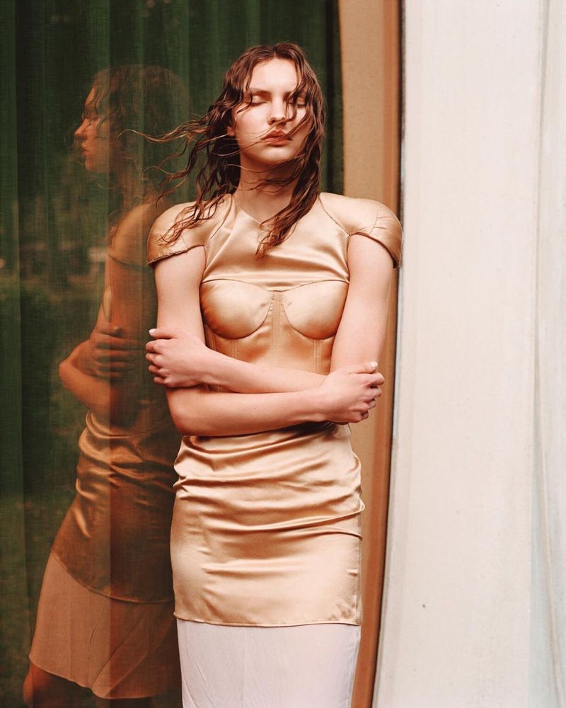 A young woman with wet hair stands with arms crossed, wearing a shiny gold top and skirt in front of a window and light-colored wall.