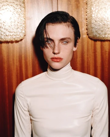 Model with short slicked dark hair wearing a white high-neck top in front of wood wall paneling and textured glass sconces.
