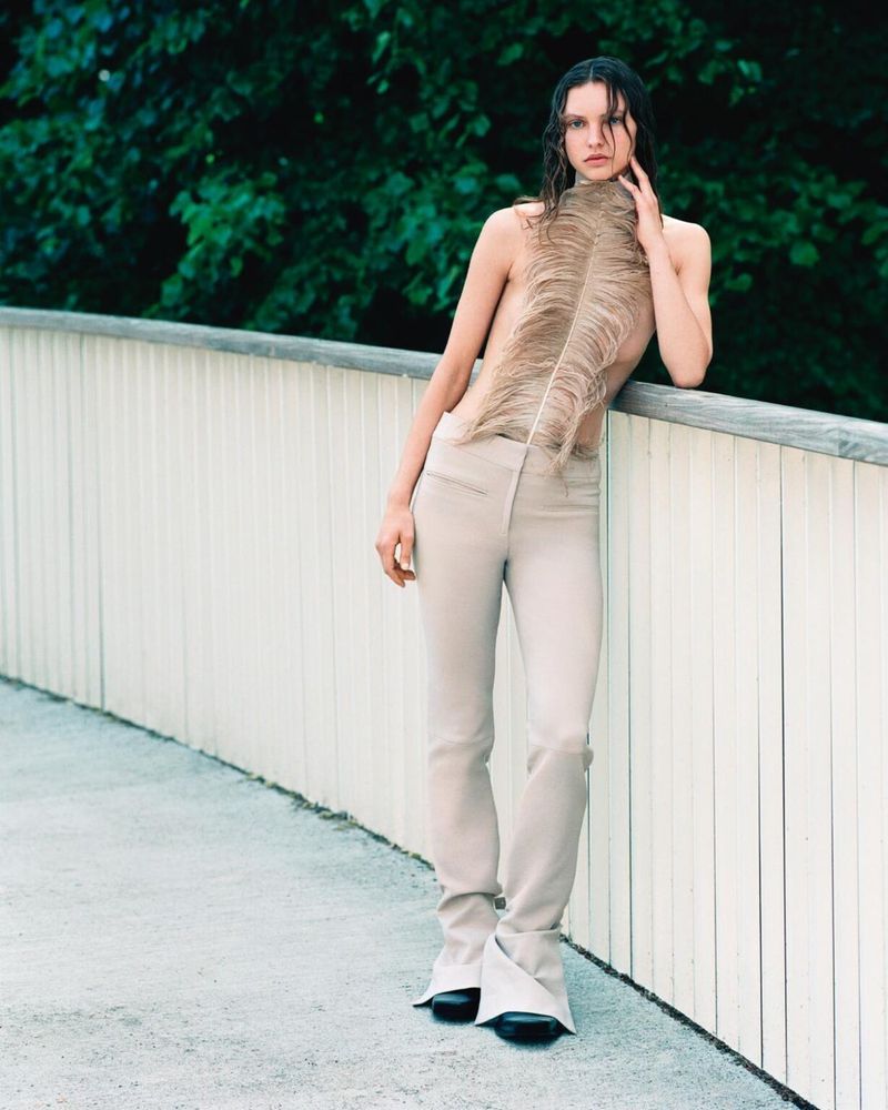 A woman in beige pants and a large feather stands on a concrete path, leaning on a white fence with a green, leafy background.