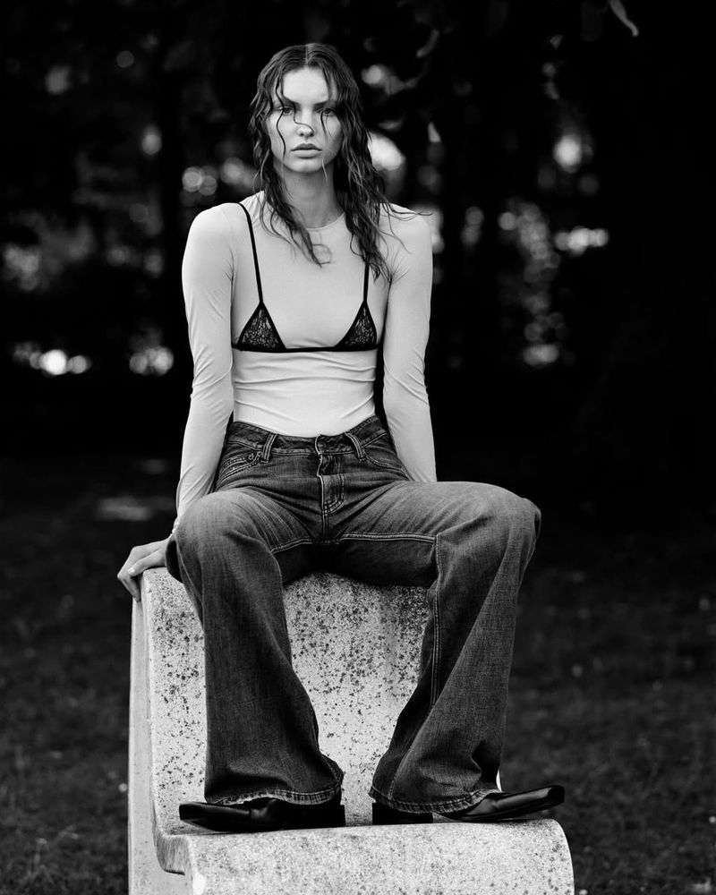 A black and white studio shot shows an adult woman with wet hair and dark jeans, sitting on a stone bench in front of blurred trees.