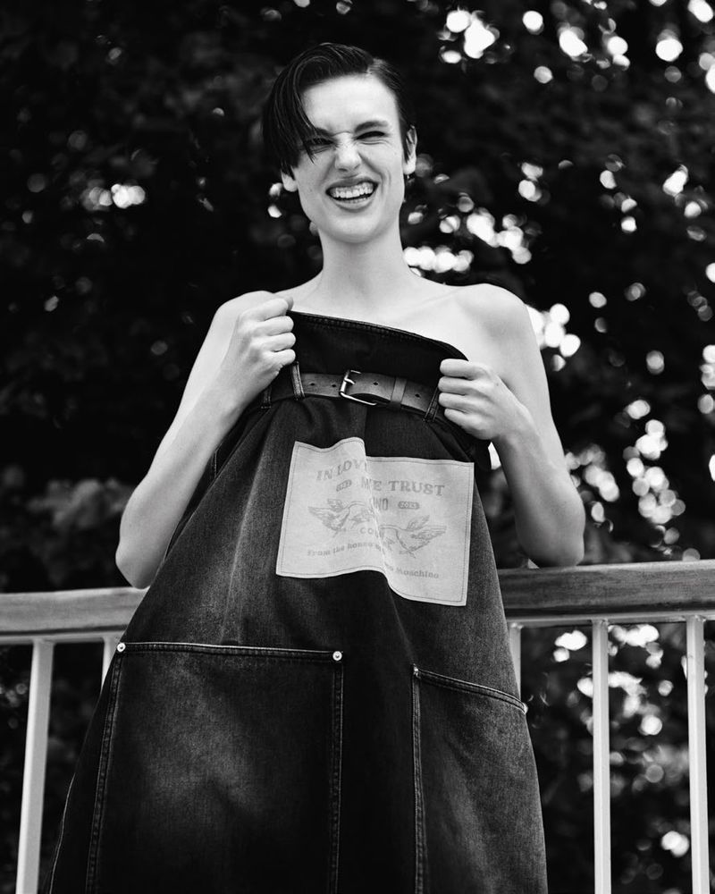 Black and white portrait of a woman smiling while holding up a dark denim apron against a railing.