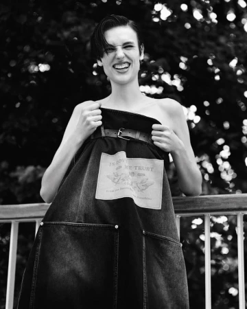 Black and white portrait of a woman smiling while holding up a dark denim apron against a railing.