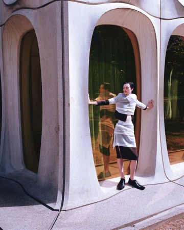 Model wearing wrapped layered garments standing within an architectural facade featuring curved concrete pillars