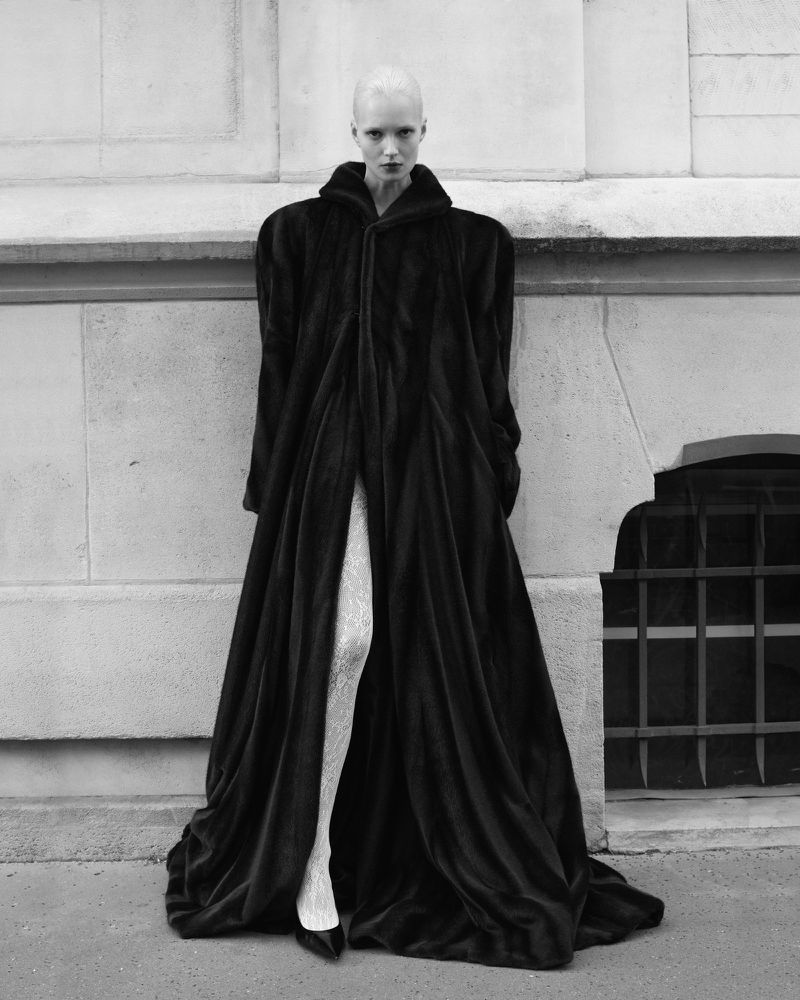 A monochrome fashion shot of a fair-skinned adult woman with shaved head wearing a long black coat and patterned tights, outdoors.