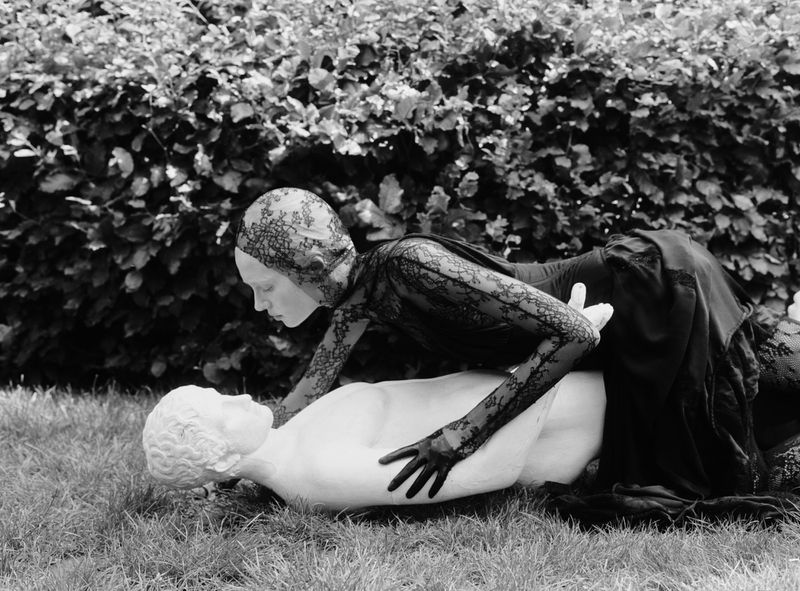 Monochrome fashion shot of a woman in lace kneeling over a sculpture on a lawn with leafy hedges. Fashion, fine art, and outdoor setting.