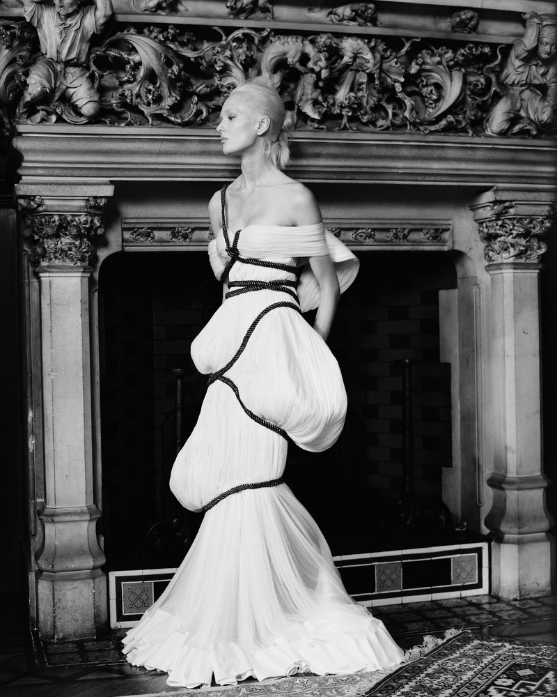 Elegant fashion shot featuring a pale-skinned model in a sculptural white gown posing in front of an ornate fireplace with carved details.