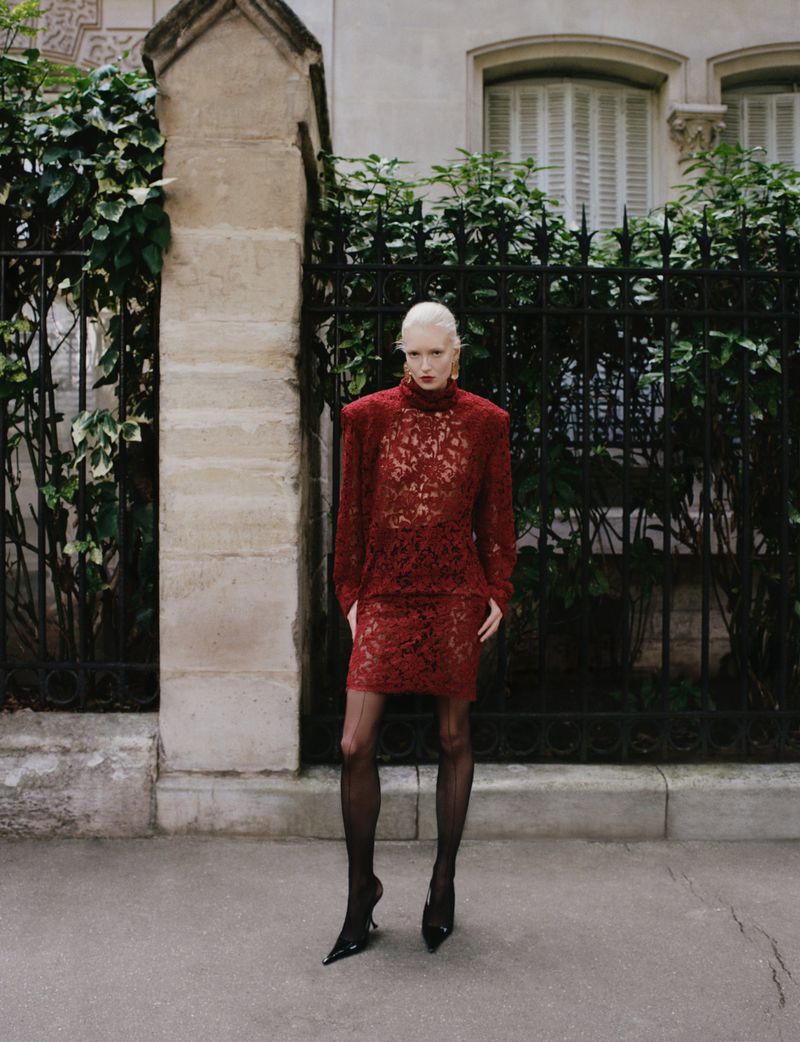 A fair-skinned woman with a blonde buzzcut poses on a city street in a red lace dress and black heels, framed by a fence and building.