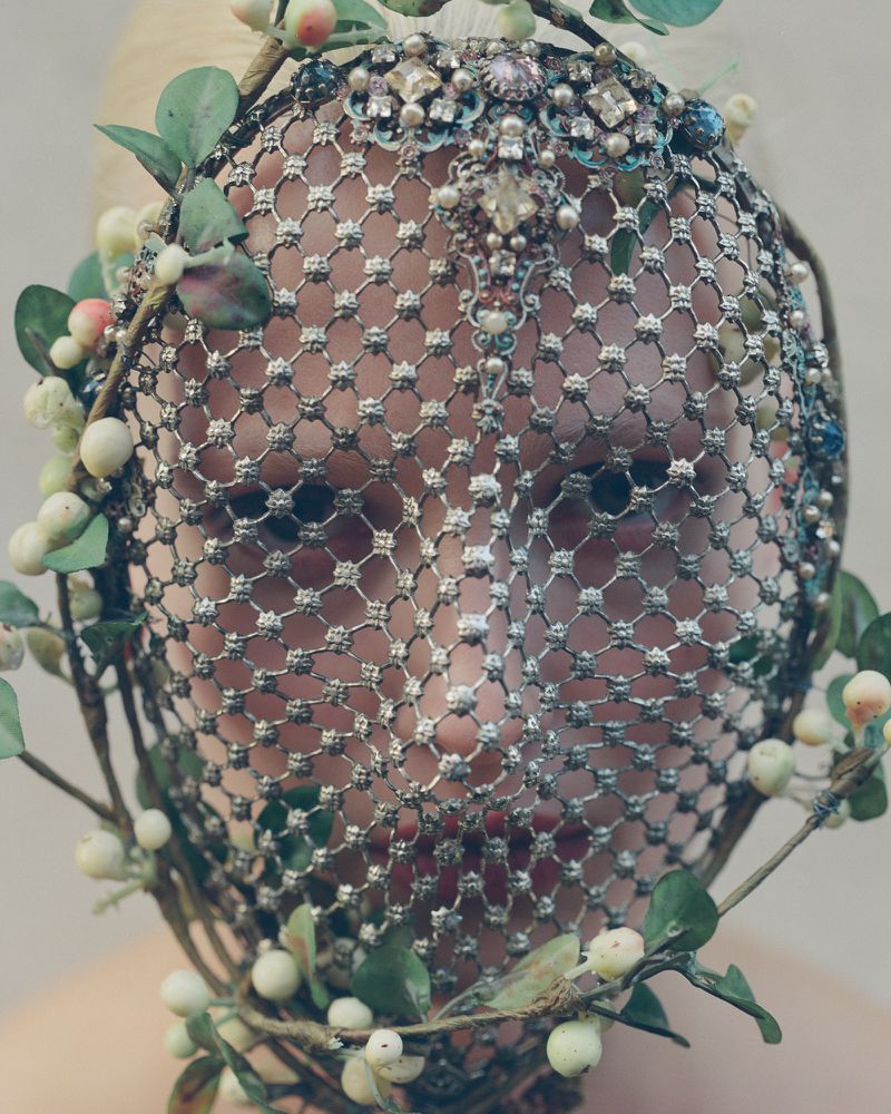 Close-up of a face covered by a decorative net, embellished with greenery and jewels, suggesting a high-fashion editorial aesthetic.