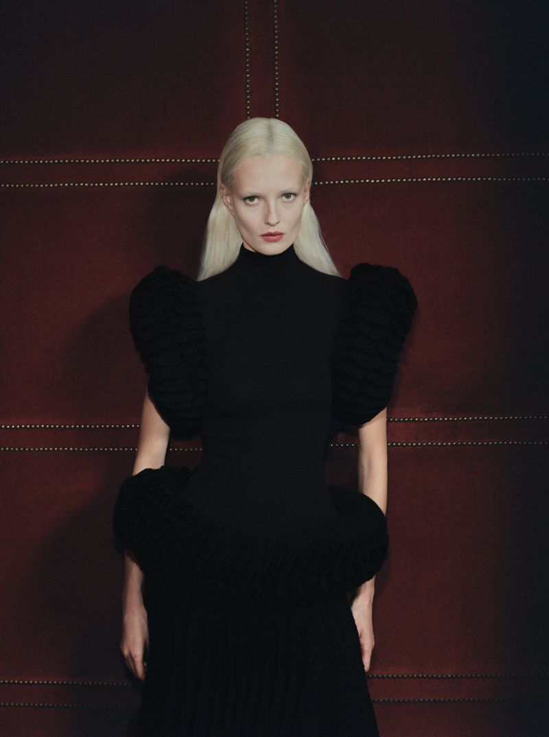 Woman in black dress with large structured shoulders posing against a red textured background with subtle linear patterns.