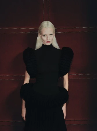 Woman in black dress with large structured shoulders posing against a red textured background with subtle linear patterns.