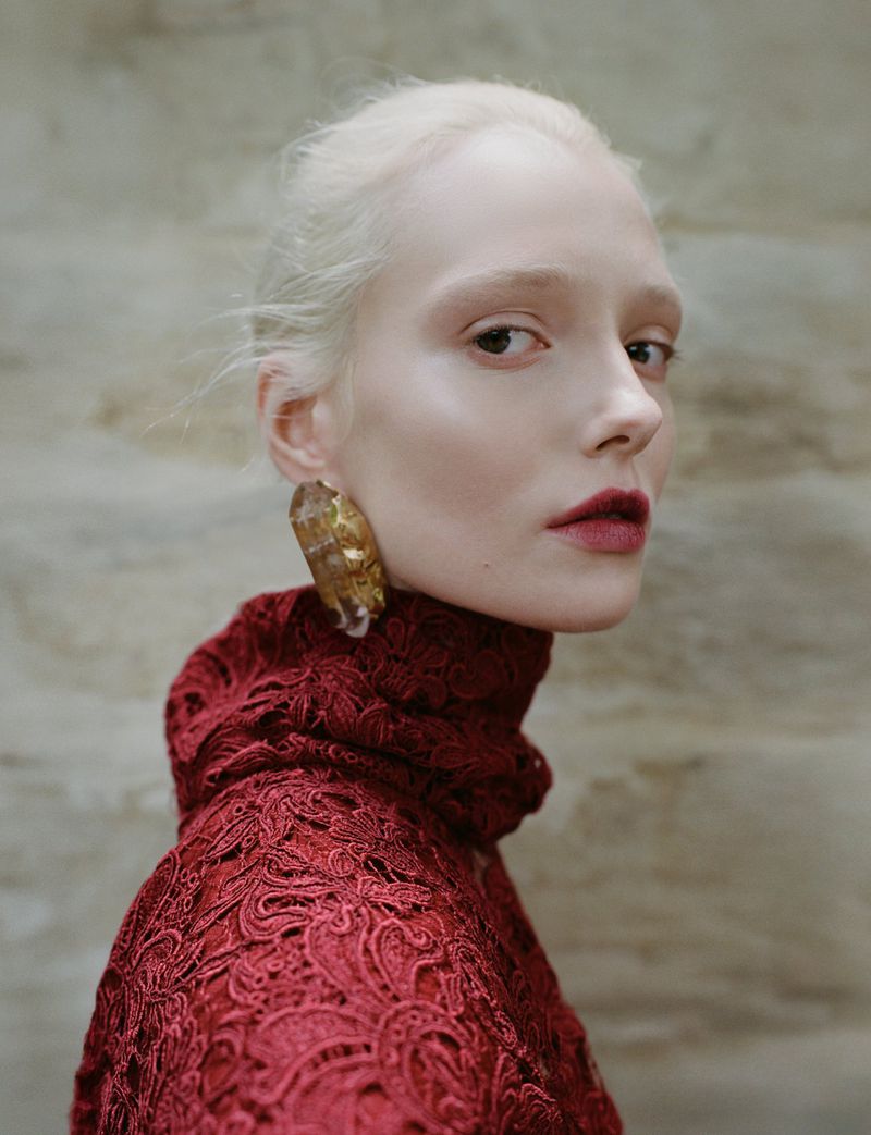 Close-up of a light-skinned woman with platinum blonde hair and dark red lipstick, wearing a red lace dress and statement earrings.