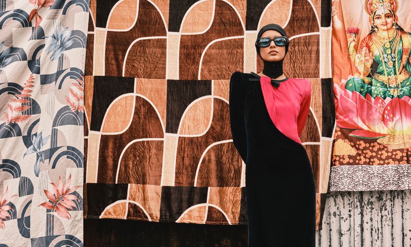 Adult female poses in a black and pink dress, wearing sunglasses and a head covering, standing in front of patterned fabric backdrops.