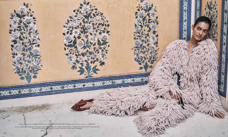 An adult female model wearing a fringed jacket reclines against a wall with floral art in a fashion editorial shot indoors.
