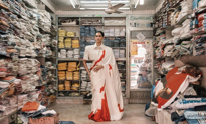 Woman in a sari poses in a clothing shop surrounded by stacks of garments and shelves filled with textiles and folded fabrics.