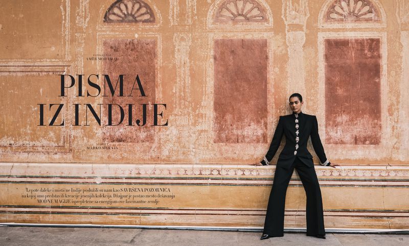 Fashion editorial shot of an adult woman in a black suit posing against an orange painted wall with architectural details in natural light.