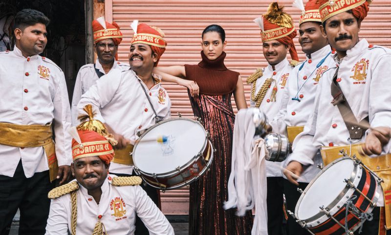Fashion shot of a slender Indian woman in a shimmering dress posing with a group of Indian musicians wearing traditional uniforms and turbans.