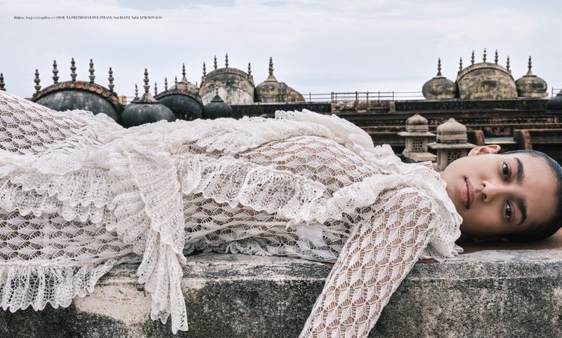 A model in a white lace dress poses on a rooftop with old domes in the background, photographed from a low angle on an overcast day.