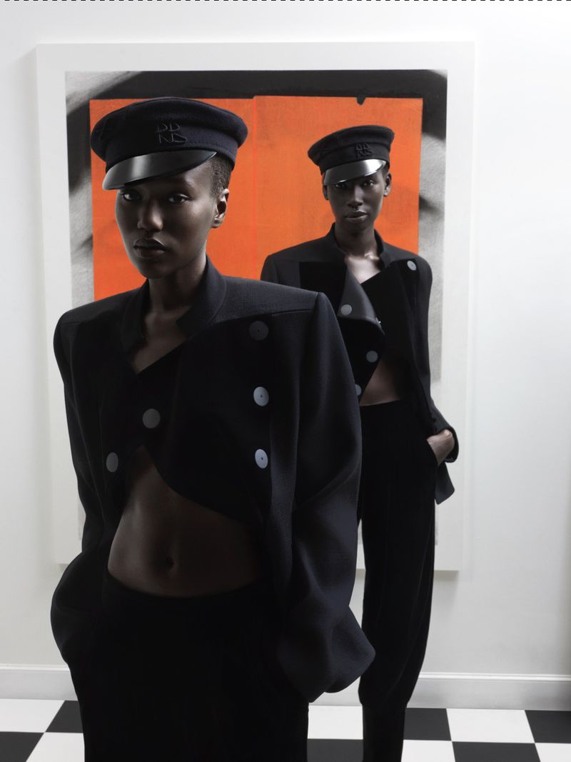 Two Black female models in black uniforms with caps posing in front of a white wall with orange abstract art on a black and white checkered floor.