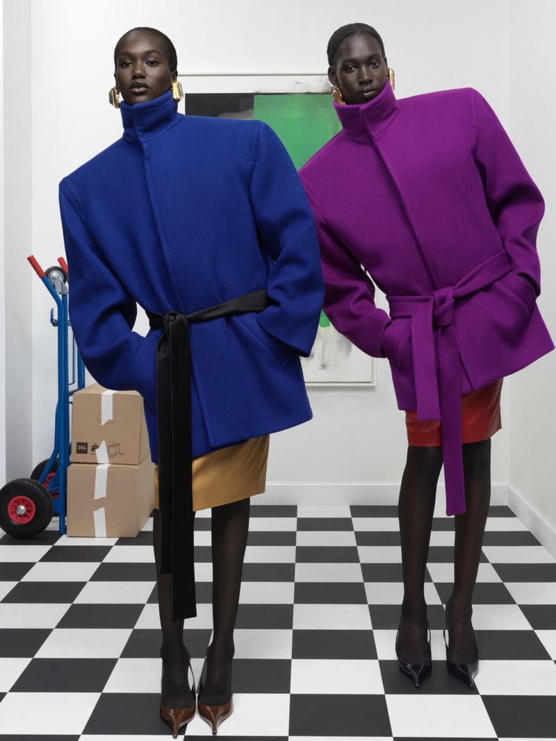 Two dark-skinned fashion models pose indoors wearing vibrant blue and purple belted coats, gold skirts, black tights, and pointed-toe heels.