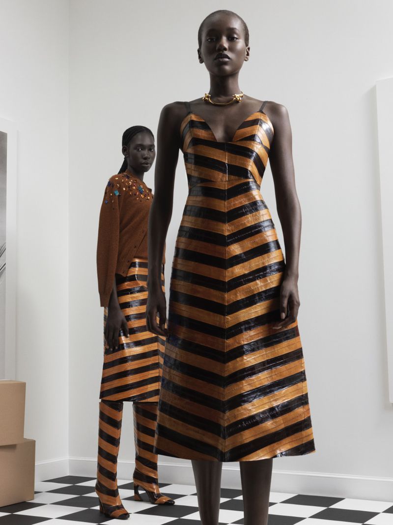 Two African-American women model brown and black striped fashion designs indoors with a checkerboard floor and minimalist decor.