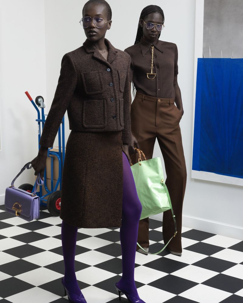 Two dark-skinned adult models wearing glasses, brown coordinated outfits, and purses stand on a black-and-white checkered floor in a studio.