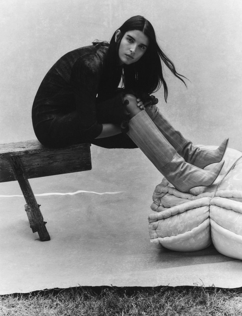 A woman with long dark hair wearing boots sitting on a wooden bench next to a large squash in a black and white fashion shot.