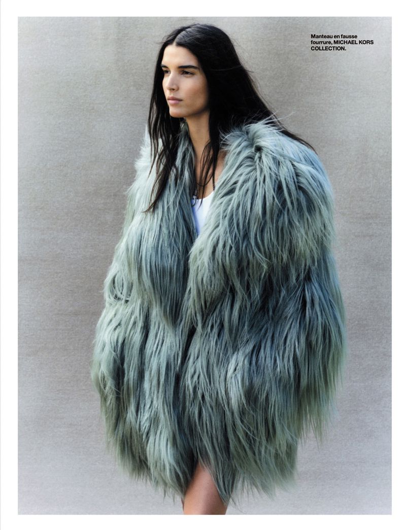 Woman in a light-blue faux-fur coat looks left against a neutral grey background in a fashion editorial, outdoor setting.