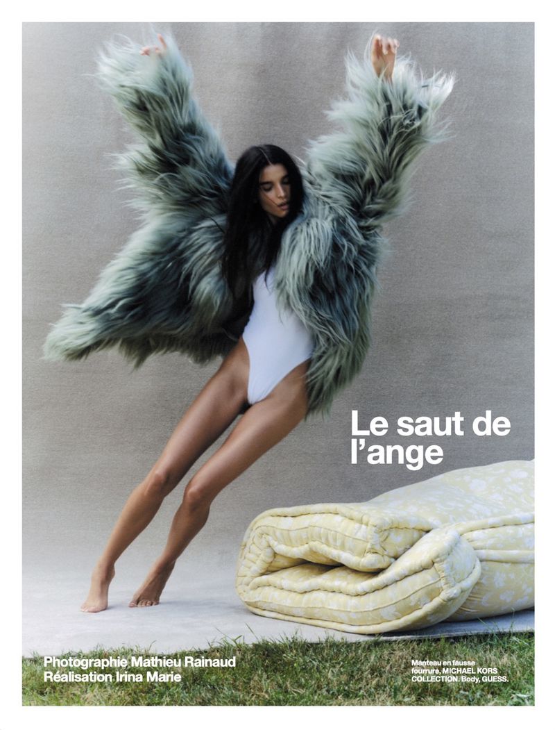 A woman in a white bodysuit and large furry coat poses next to a rolled mattress on a neutral background, bathed in natural light.
