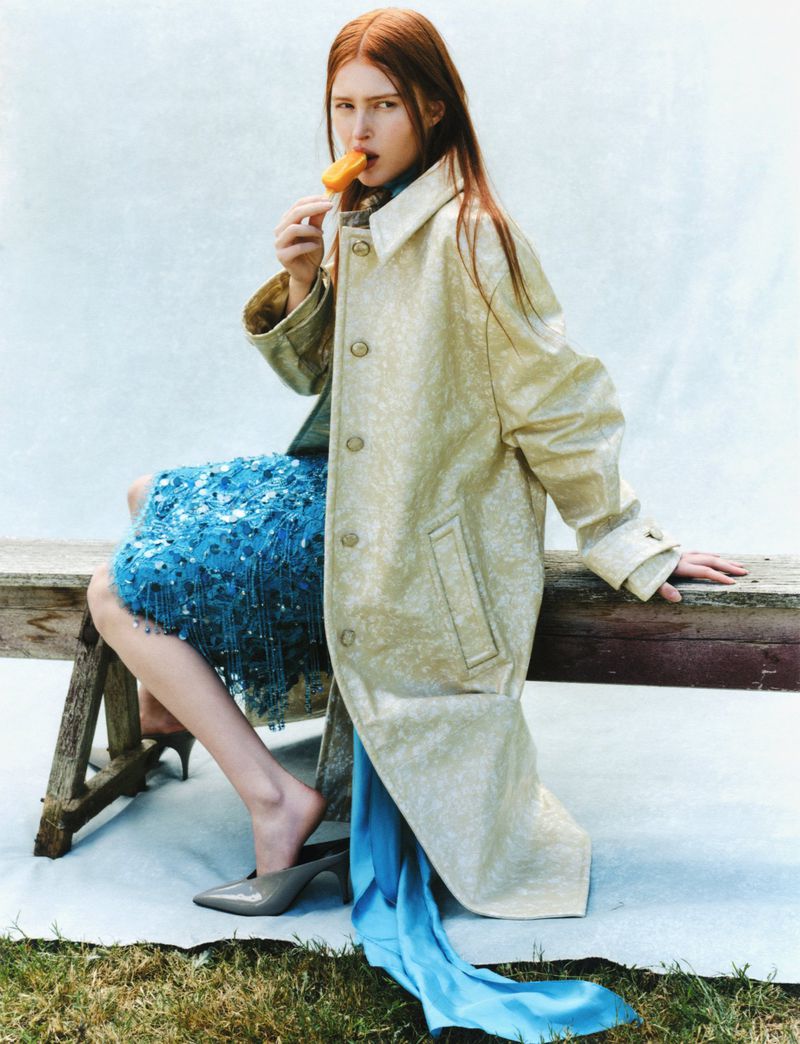 A red-haired woman in a tan coat sits on a bench outdoors eating a yellow popsicle. She wears a blue skirt with a draped blue scarf.