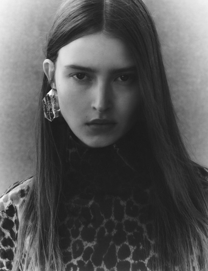 Portrait of a woman with long brown hair wearing an animal-print top and statement earring in a monochrome photo studio setting.