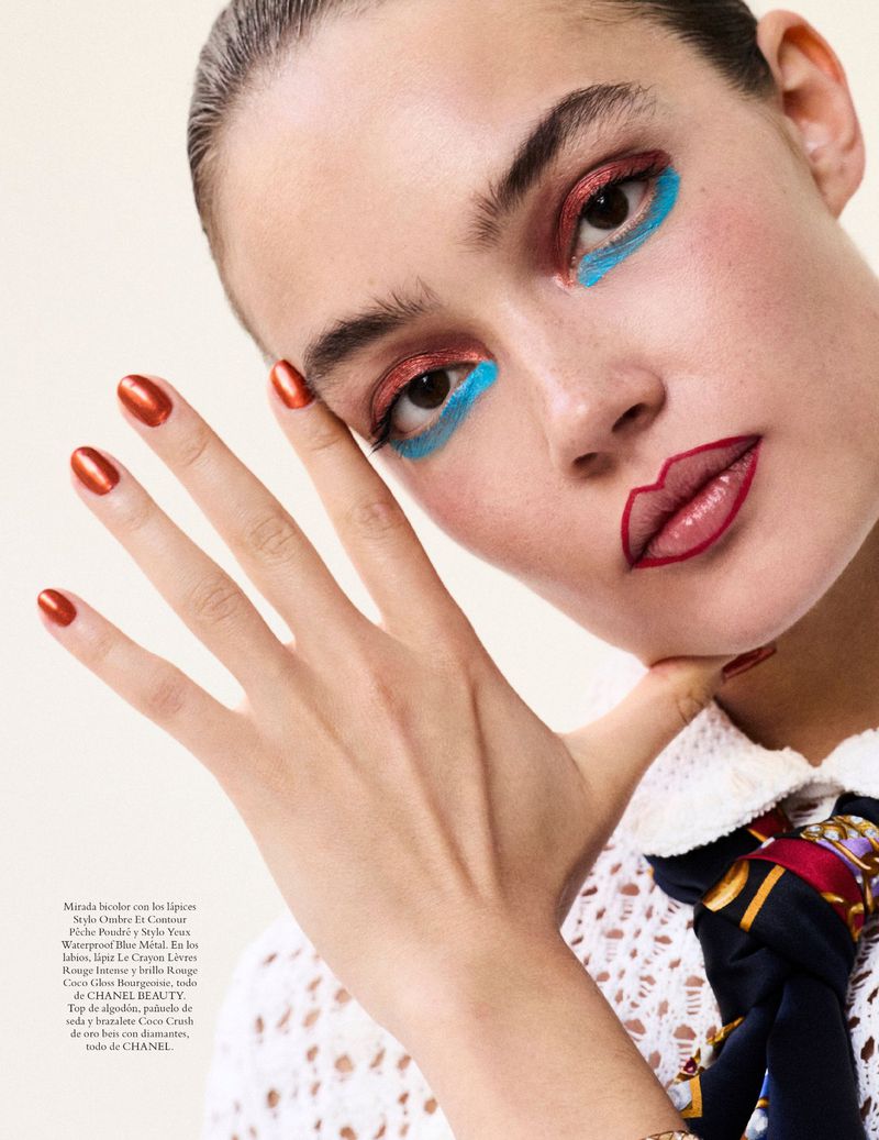 Close-up of a woman with colorful makeup, including red lipstick outlines, blue eyeliner, and shimmering copper eyeshadow, posing indoors.