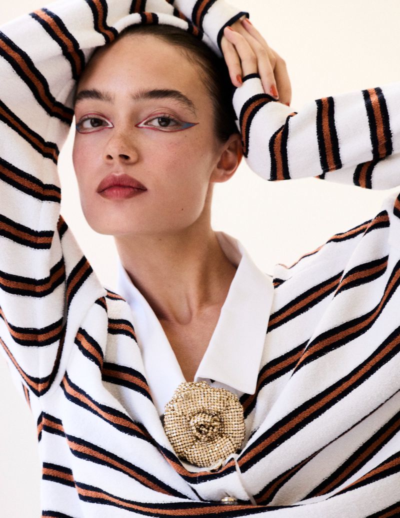 Portrait of an adult female wearing a striped sweater and a jeweled brooch, indoors in front of a light coloured background.
