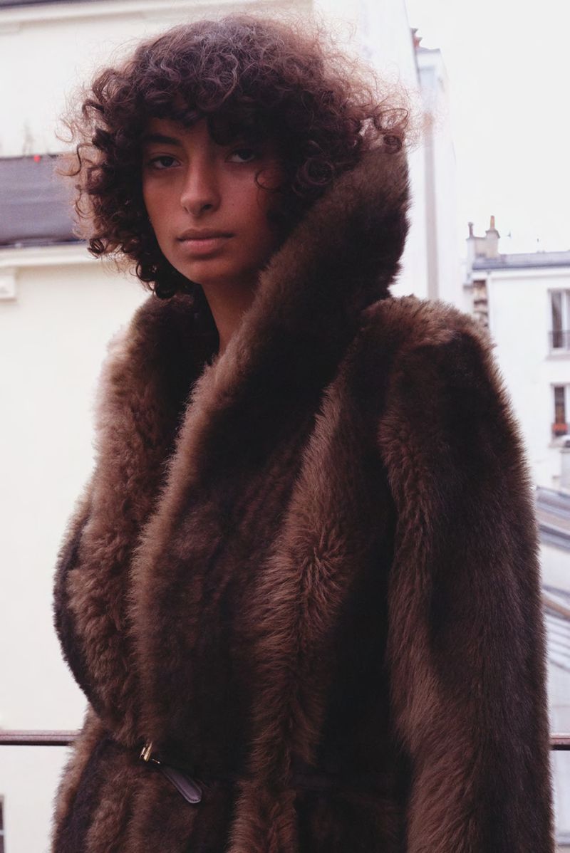 Model with curly dark hair wearing a fluffy brown winter coat against an outdoor building background